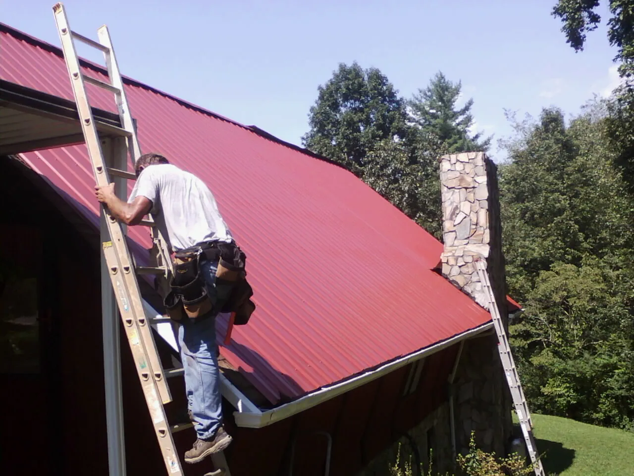 Roofer on a ladder beside a red metal roof and stone chimney — Soffit & Fascia Repair by Roofing Brothers in Bird In Hand